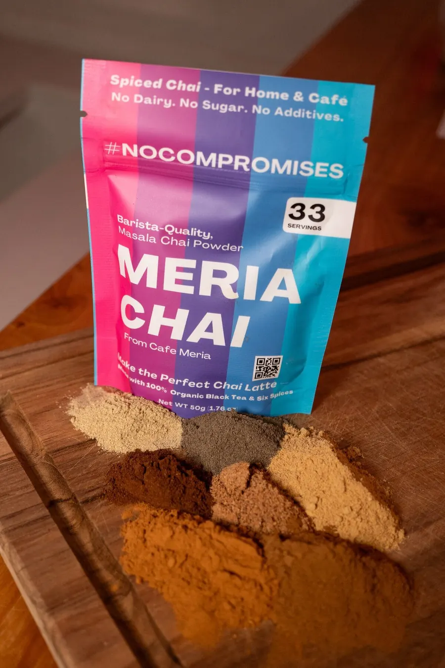 A Meria Chai 50 g pouch standing on a wooden board, with the spices in small piles in front of it
